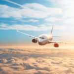 Photograph of a plane flying in the air over clouds with the sun to it's left.