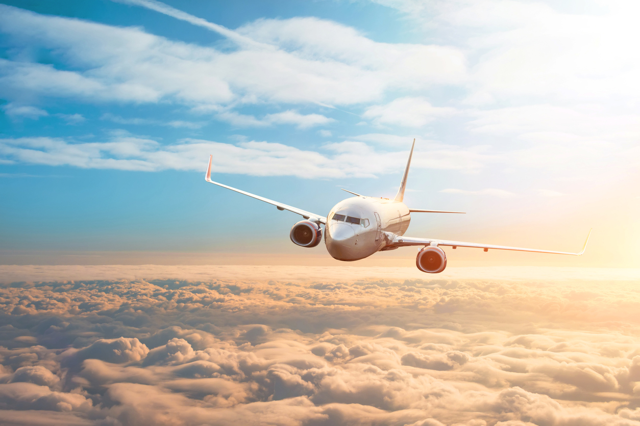 Photograph of a plane flying in the air over clouds with the sun to it's left.