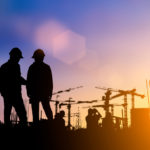 silhouette of two construction workers standing in front of a construction site with a purple, yellow sky.