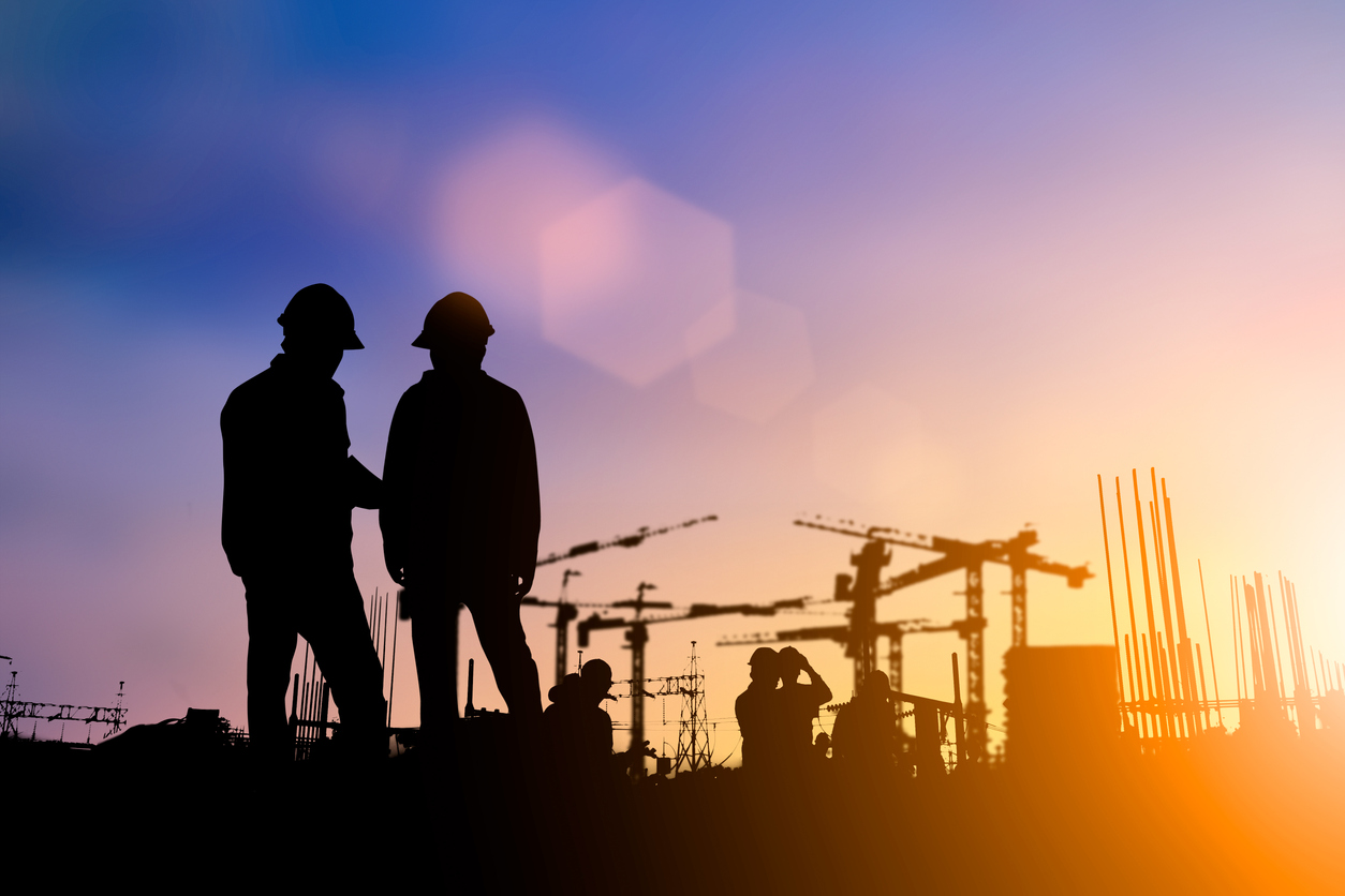 silhouette of two construction workers standing in front of a construction site with a purple, yellow sky.
