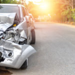 Photograph of a silver car that's been in a car accident on a road surrounded by trees.