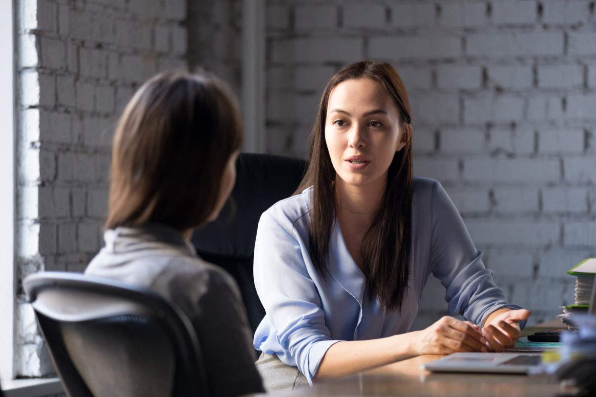 Female professional wearing a button up shirt talking to another woman in an office.