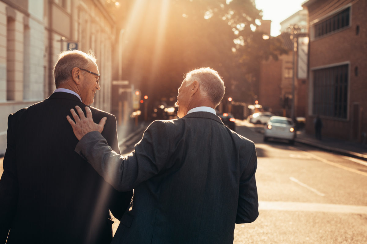 Two older men walking down the street at sunset smiling at each other.