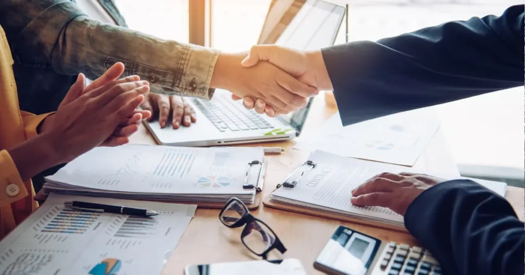 Close-up of business professionals shaking hands over contract documents and financial charts, symbolizing a real estate joint venture agreement and partnership.